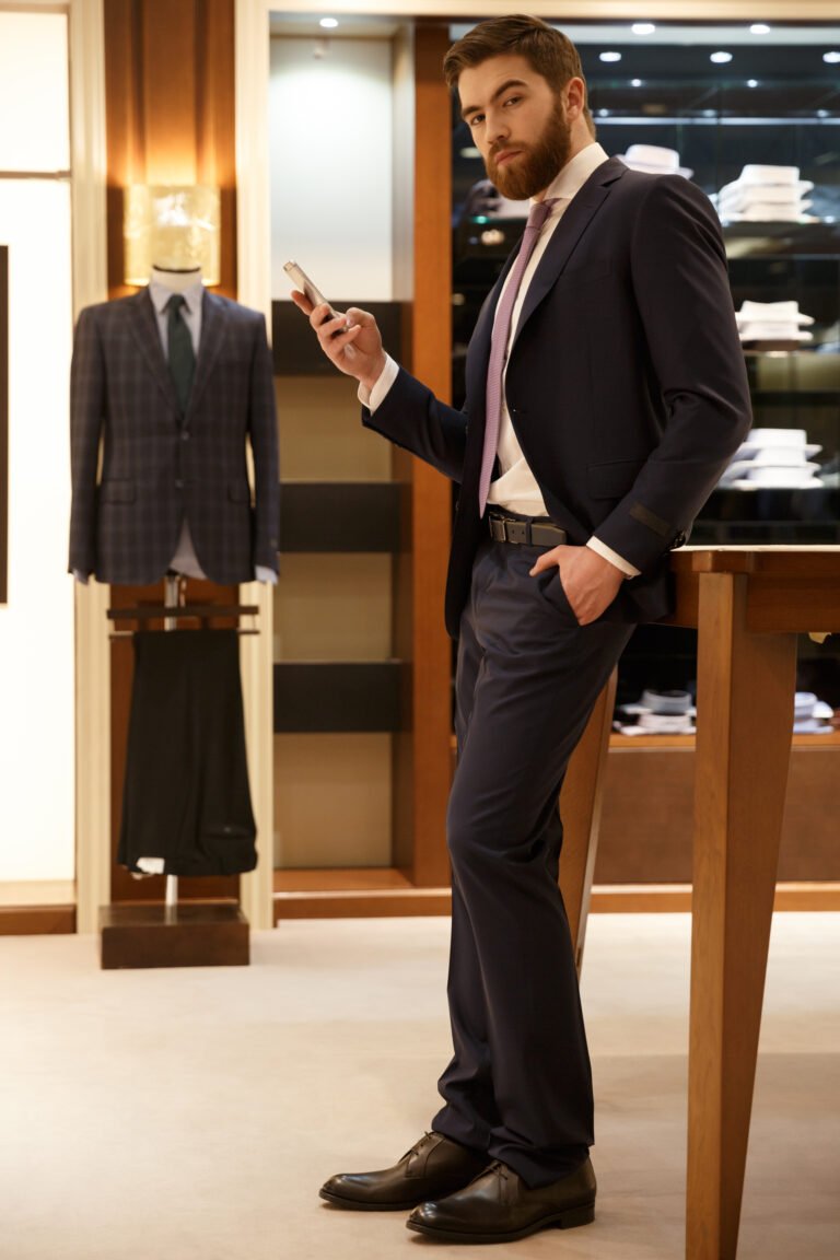 Vertical image of bearded man in suit  holding smartphone in his hand and holding one arm in pocket while looking at camera. Full length portrait